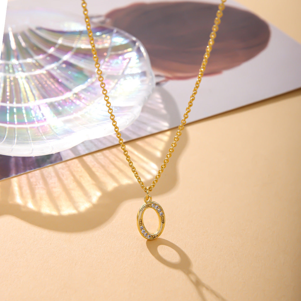 The K.Bola Fashion English 26 Letters Inlaid Zircon Copper Necklace is elegantly showcased on a smooth beige surface, featuring a stunning oval pendant. In the background, a geometric glass decoration adds colorful reflections.