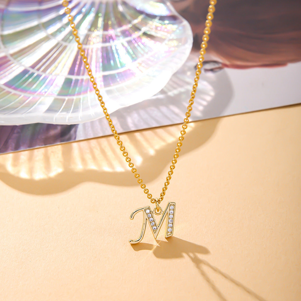 Displayed on a peach surface, the K.Bola Fashion English 26 Letters Inlaid Zircon Copper Necklace, an elegant gold O-shaped chain womens piece, showcases a partially diamond-encrusted M pendant. A colorful shell and magazine enhance its presentation.