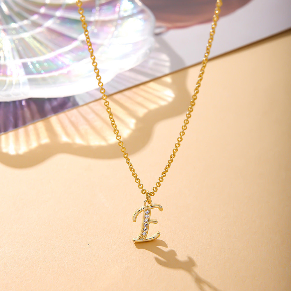 The K.Bola Fashion English Necklace, crafted from an alloy featuring an O-shaped chain and E shaped pendant with zircon inlay, rests on a light beige surface beside a colorful iridescent shell.