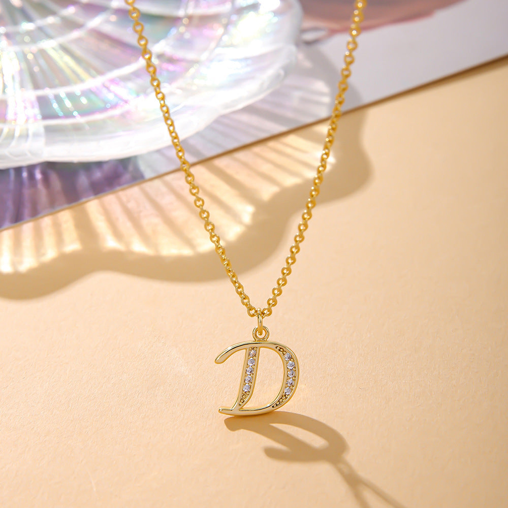 The K.Bola Fashion English 26 Letters Inlaid Zircon Copper Necklace features a stunning D pendant adorned with crystals on an O-shaped chain, elegantly displayed on a beige surface with an iridescent dish peeking through.