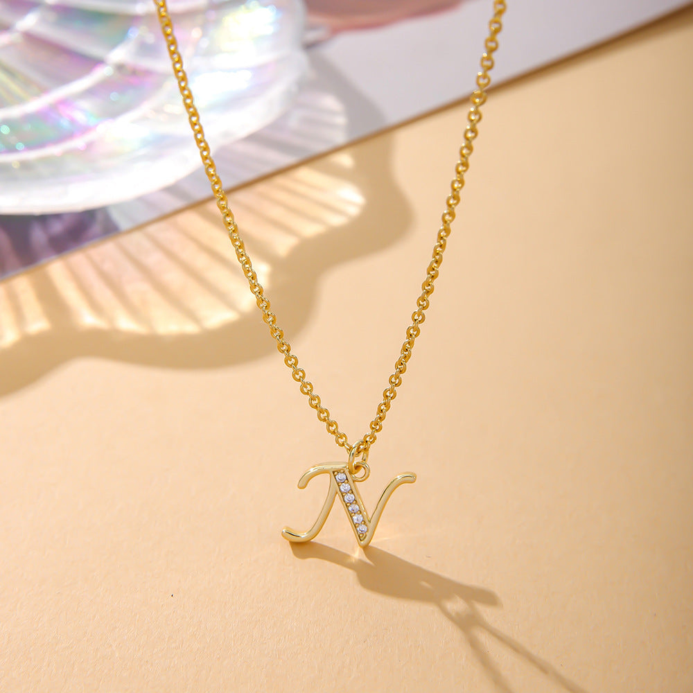 A K.Bola necklace from the Fashion English 26 Letters Inlaid Zircon Copper line showcases an N pendant with gemstones on a gold O-shaped chain, elegantly laid on a soft beige surface with a subtle rainbow-reflective decorative background.