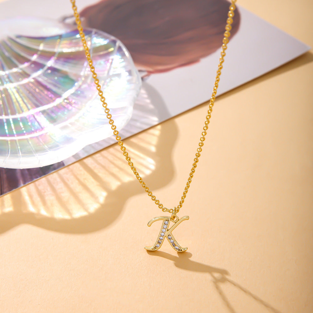 The K.Bola Womens Fashion English Letter K Necklace, featuring an O-shaped chain, shines with its inlaid zircon on a pendant of copper alloy. Resting on a beige surface next to an iridescent shell and blurred photo, the pendant gleams in the soft natural light.