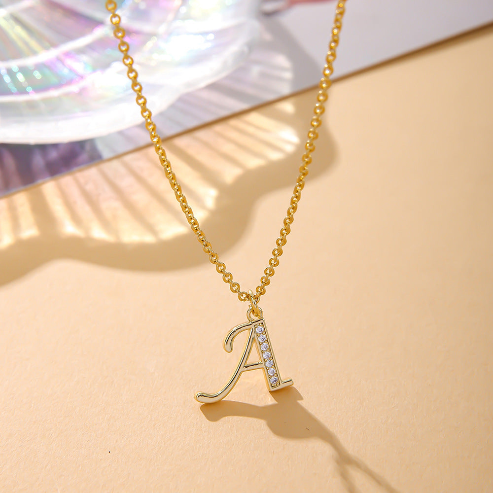 On a peach surface, the K.Bola Fashion English 26 Letters Inlaid Zircon Copper Necklace displays an elegant gold pendant with the letter A, adorned with gemstones. Its intricate design is complemented by an alloy O-shaped chain, shimmering softly against the iridescent background.