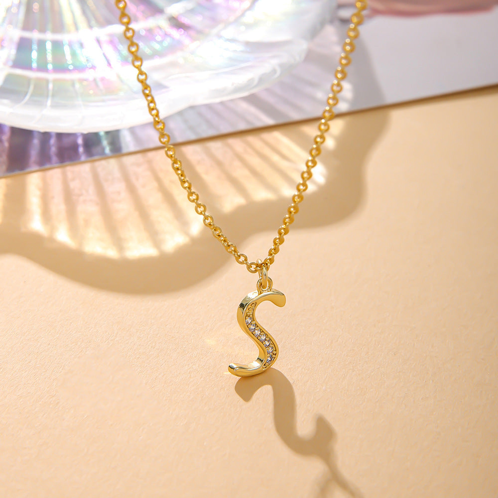 The K.Bola Fashion English 26 Letters Inlaid Zircon Copper Necklace, featuring a letter S pendant with small gemstones, rests on a light surface casting a shadow. An iridescent shell-like decor is positioned behind it.