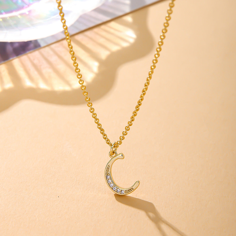 The K.Bola Fashion English 26 Letters Inlaid Zircon Copper Necklace features a gold crescent moon pendant with shimmering gemstones, elegantly resting on a light beige surface. The O-shaped chain enhances its allure, with sunlight casting soft shadows for a warm, elegant ambiance.